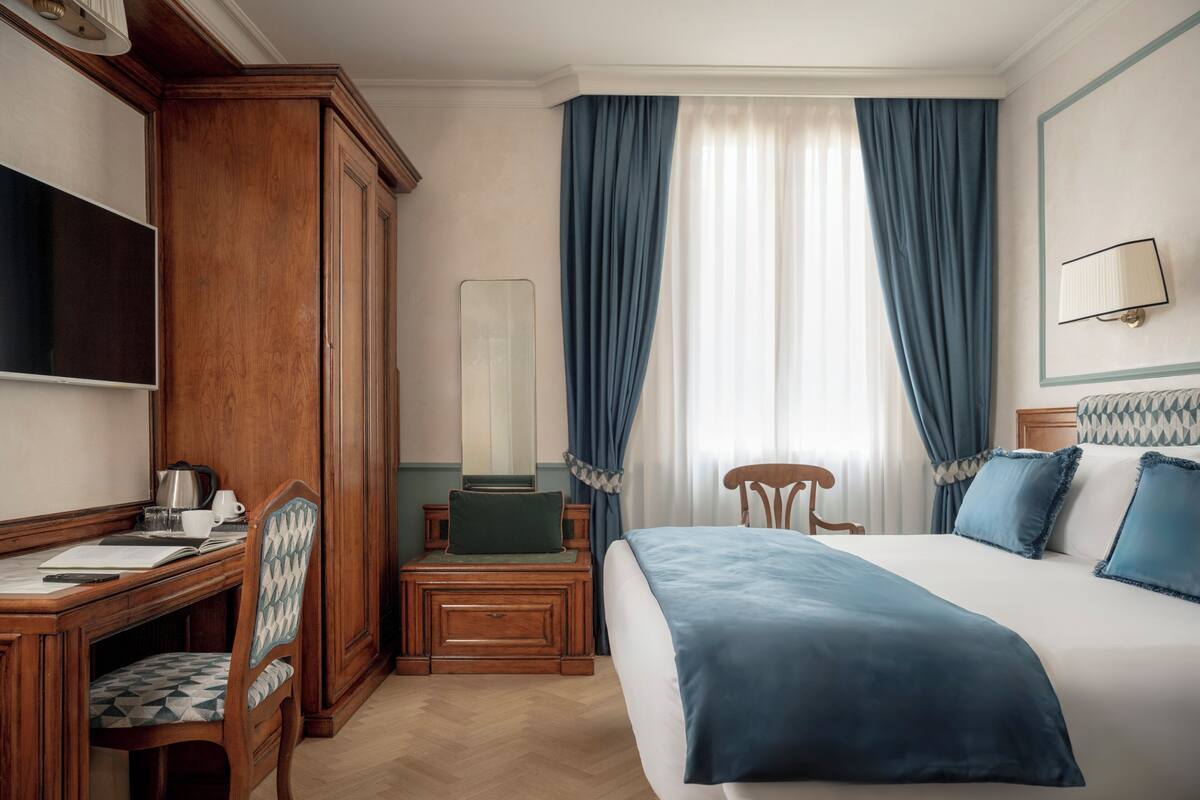 A hotel room with a bed, a desk with a chair, a wardrobe, a mirror, and a window with curtains.