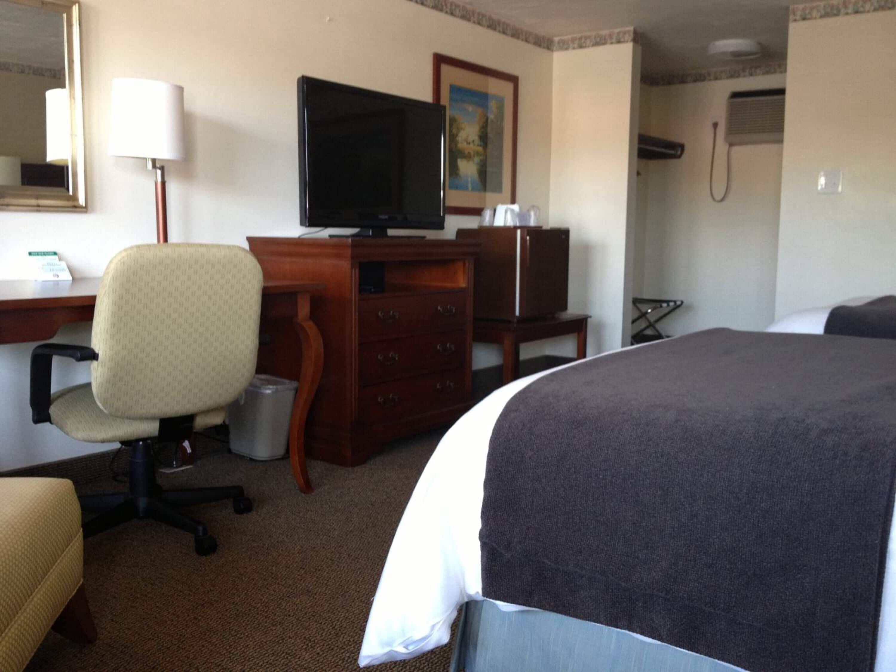 Quarto de hotel com cama, escrivaninha com computador, cadeira, televisão e um pequeno armário.