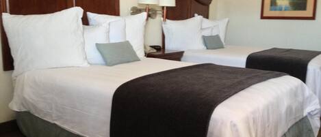 A hotel room with two beds, each with white linens and a dark throw blanket. There is a nightstand with a lamp between the beds.