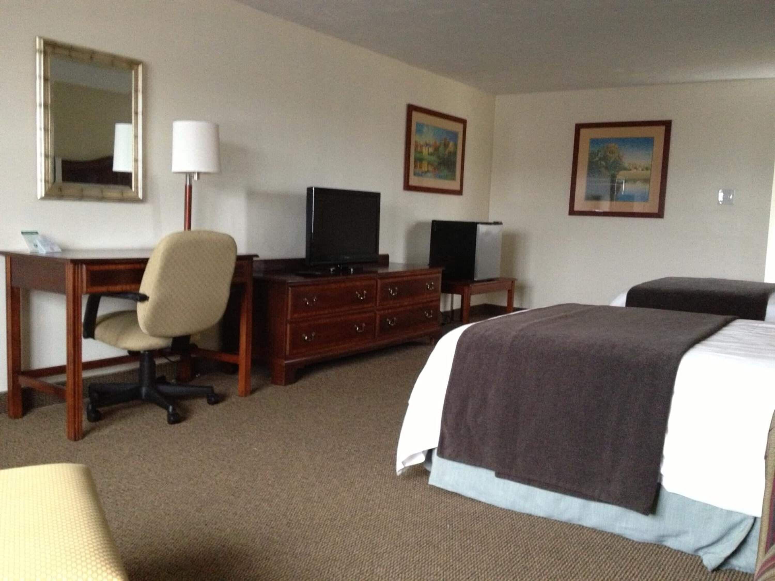 A hotel room with a bed, a desk with a chair, a television, a dresser, and two framed pictures on the wall.