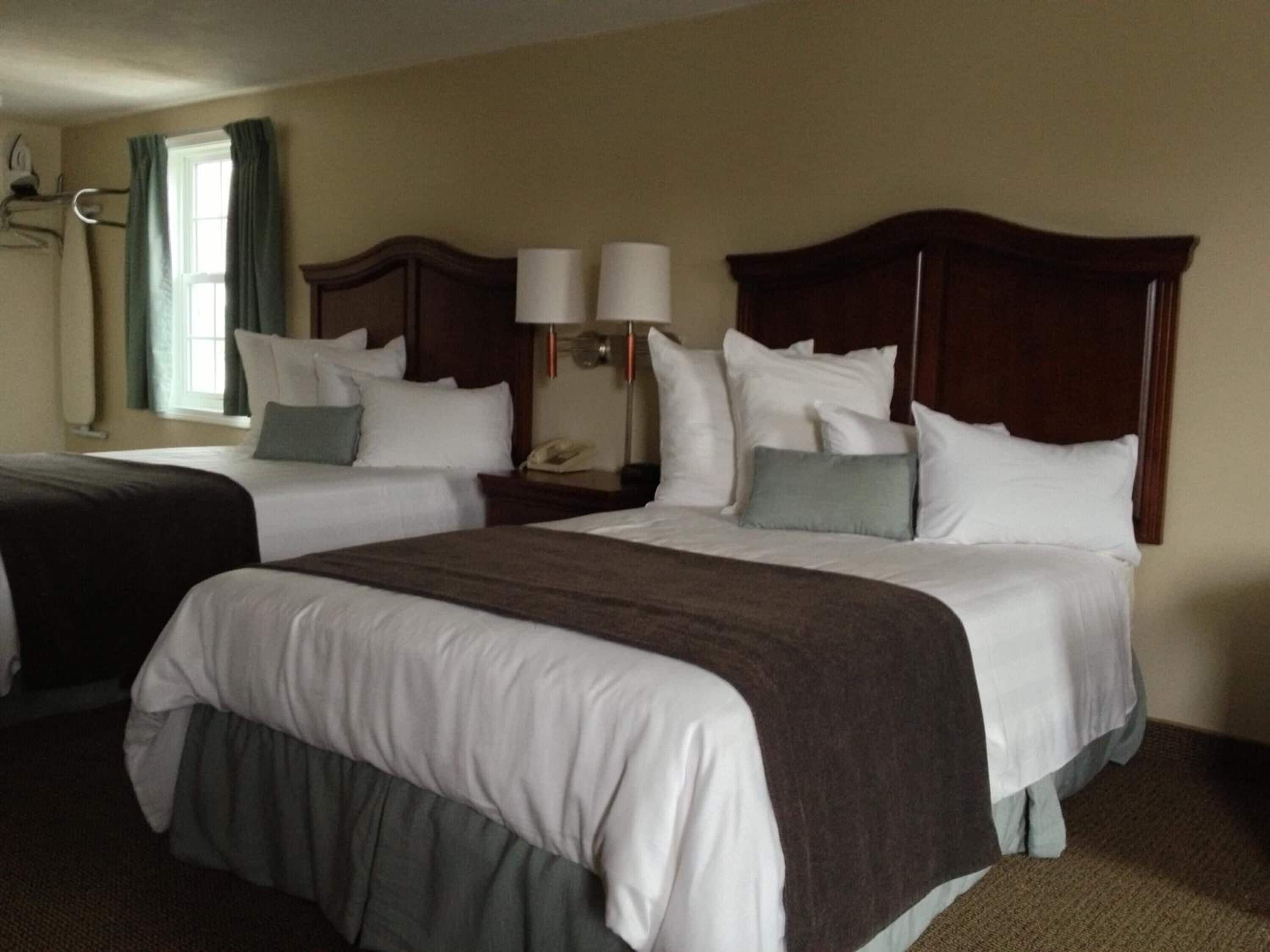 A hotel room with two beds, a nightstand with a lamp, and a window with curtains.