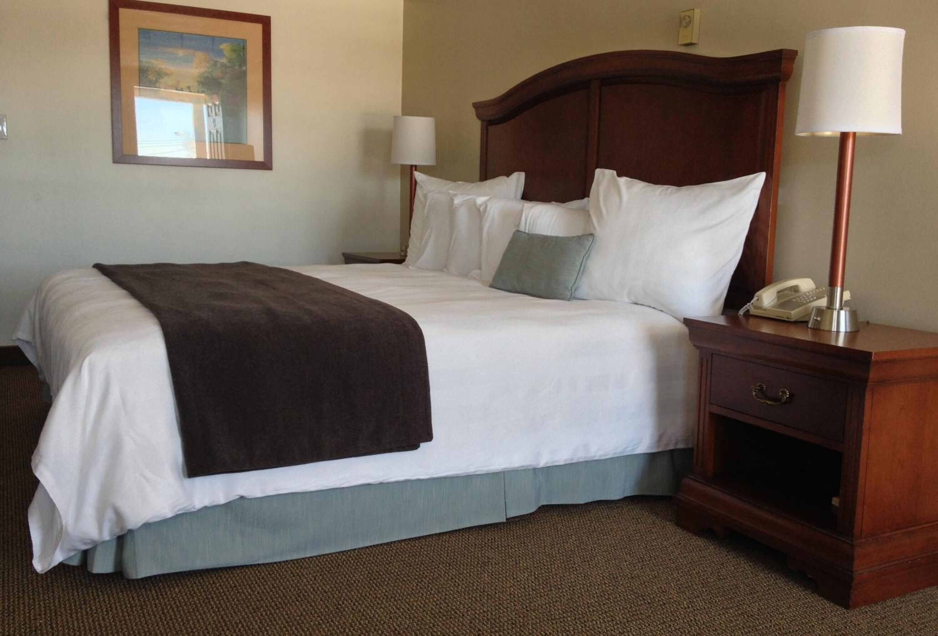 A neatly made bed with white linens, a brown throw blanket, and a wooden headboard. There is a bedside table with a lamp and a telephone.