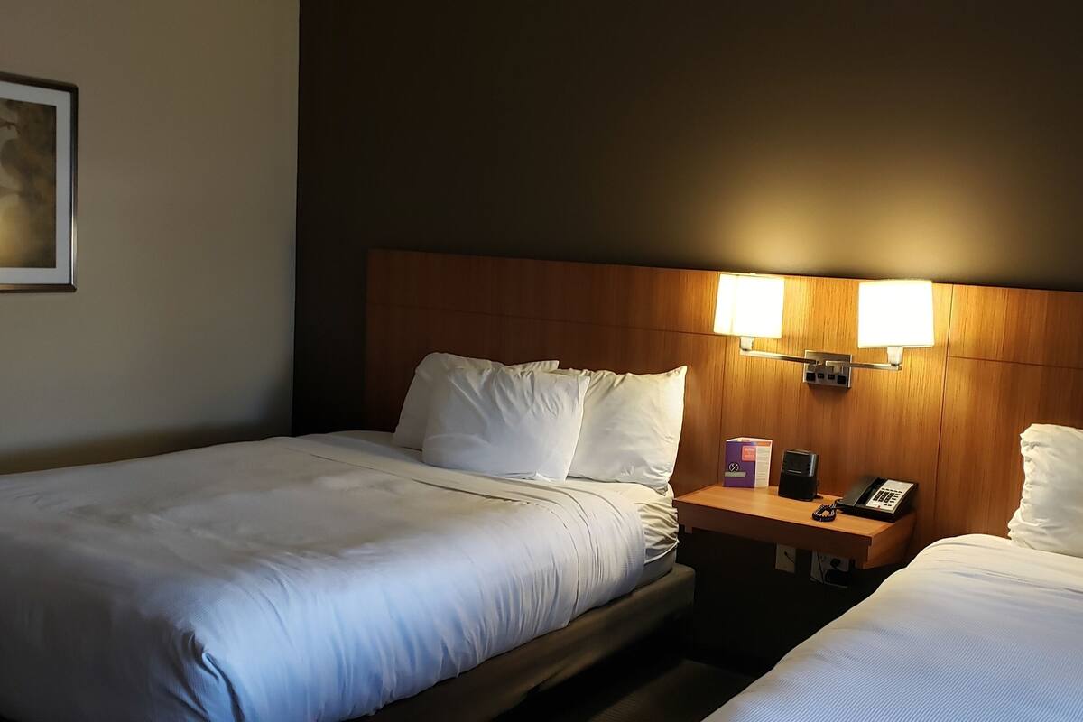 Een hotelkamer met twee bedden, een nachtkastje met een telefoon en een doos, en een wandlamp.