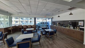 Interior de un restaurante con mesas y sillas, una zona de cocina y amplias ventanas con vistas a un estacionamiento.