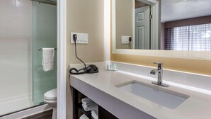 A bathroom with a glass shower enclosure, a white sink with a mirror, and a towel rack. - Baymont by Wyndham Pawleys Island (Pawleys Island)