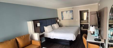 Quarto de hotel com cama, escrivaninha, cadeira, sofá e banheiro visível através de uma porta aberta.