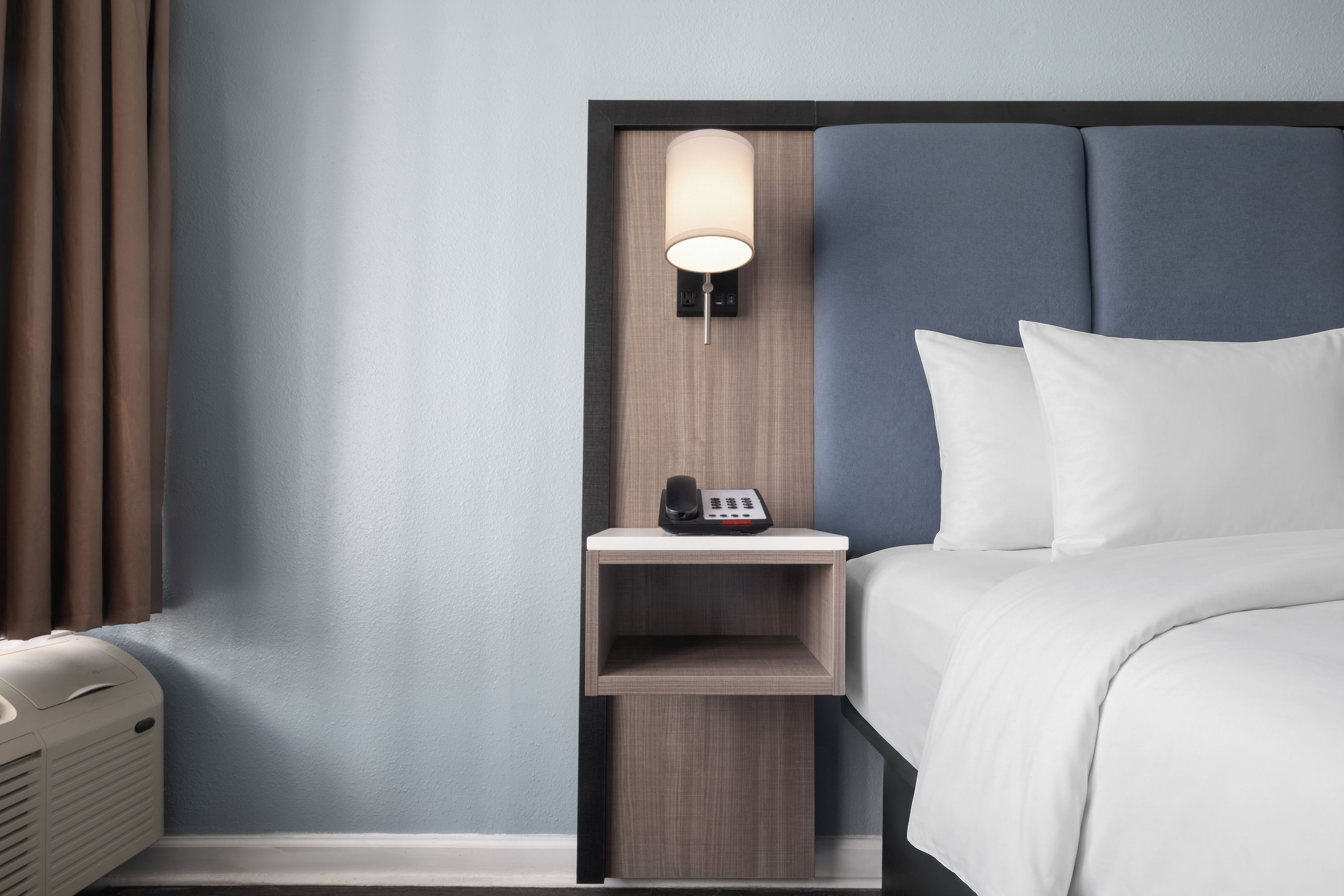 A hotel room with a bed, a bedside table with a lamp, a phone, and a wall-mounted control panel.