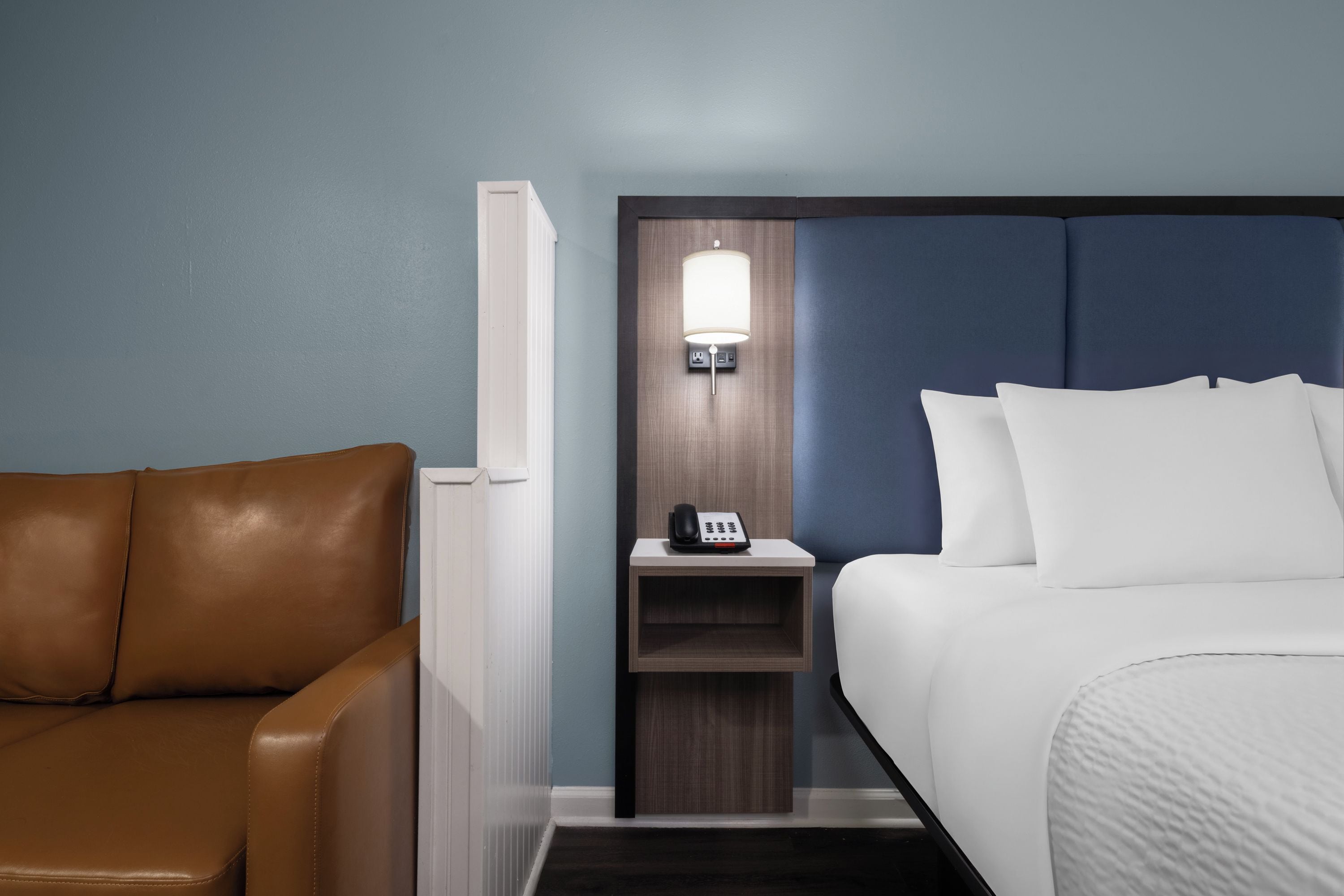 A hotel room with a brown leather chair, a bed with white bedding, a nightstand with a lamp, and a telephone.