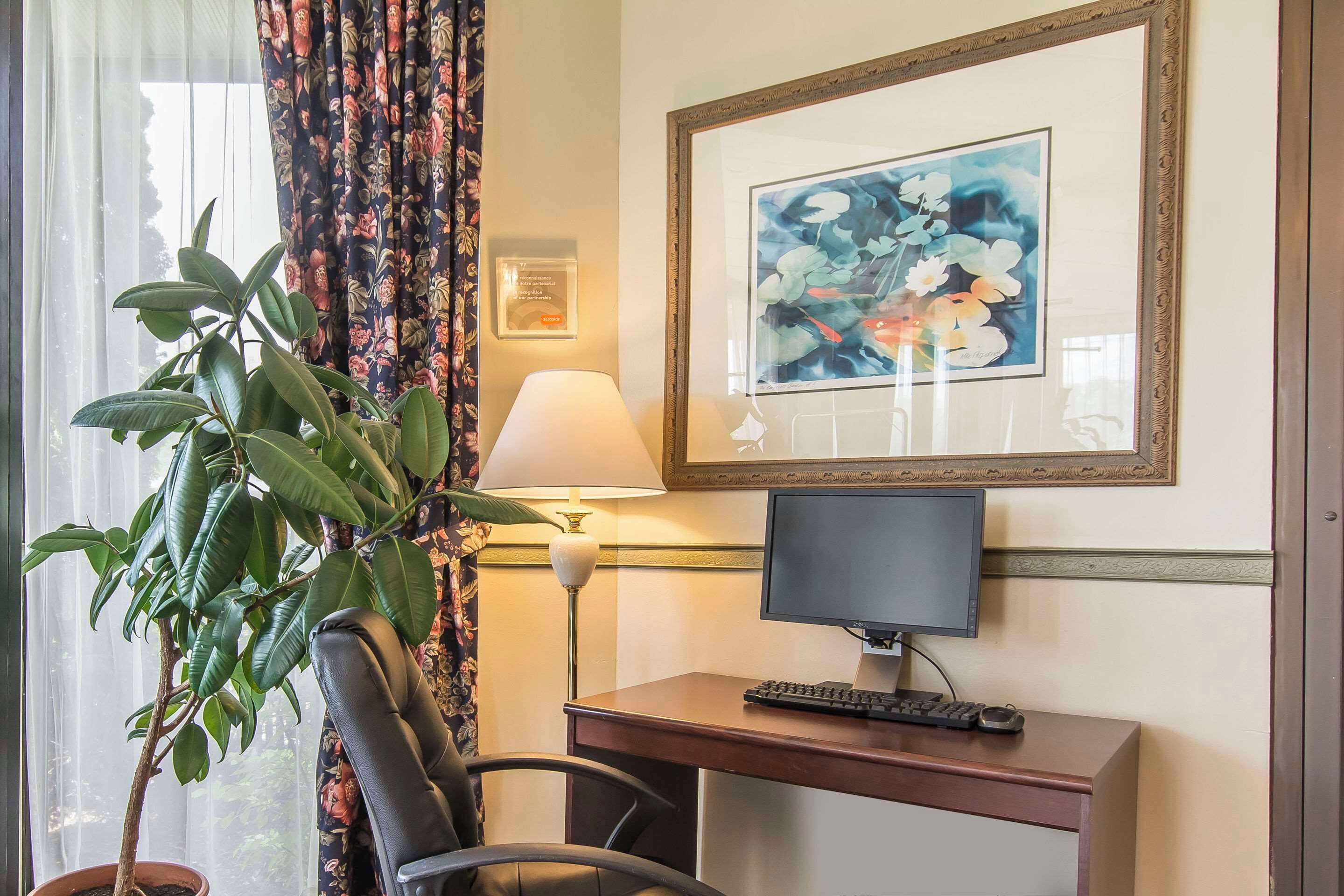 A hotel room with a desk, chair, computer, lamp, potted plant, and a framed painting on the wall.