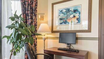 A hotel room with a desk, chair, computer, lamp, potted plant, and a framed painting on the wall.