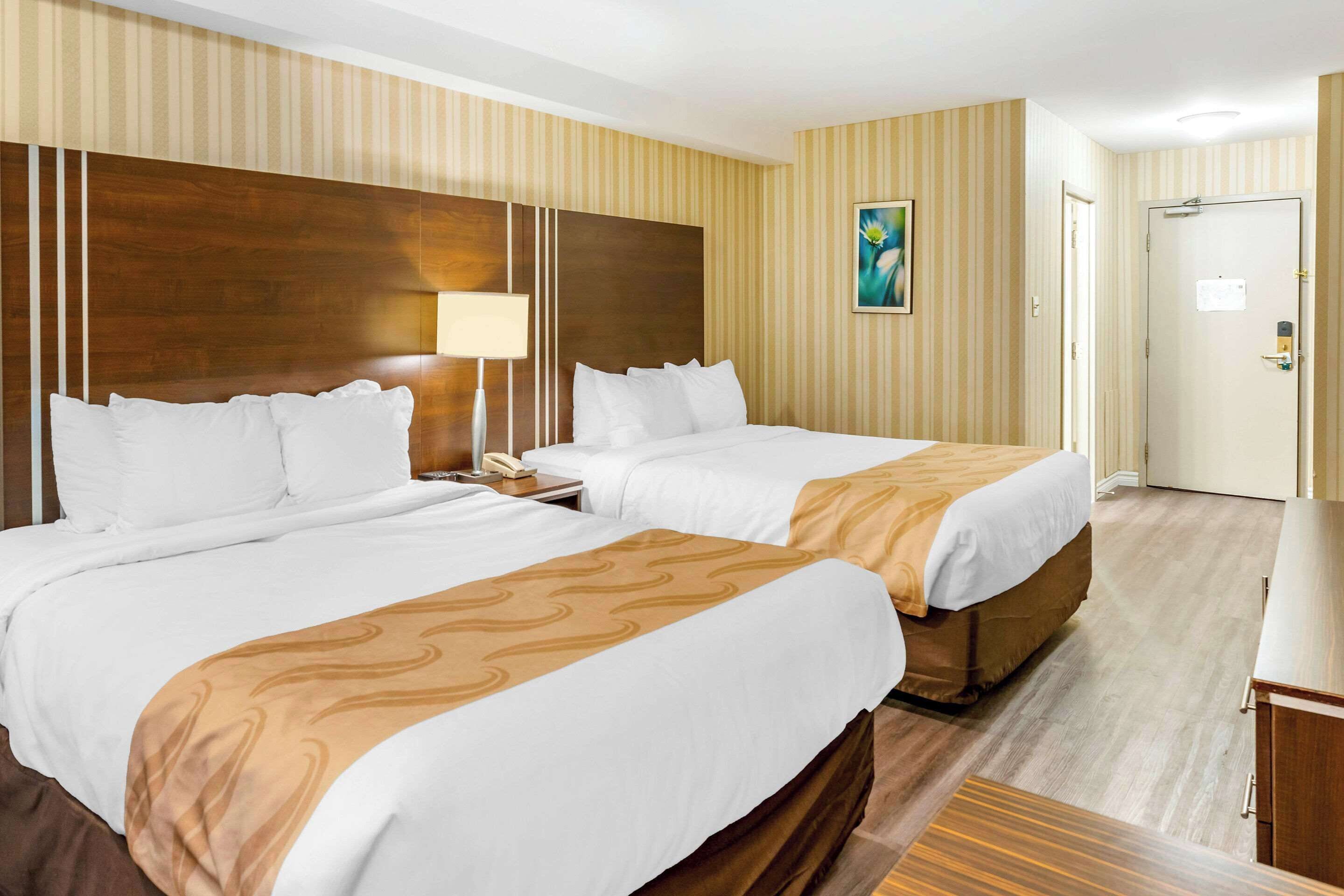 Hotellihuone, jossa on kaksi sänkyä, yöpöytä lampun kanssa, ovi ja taulu seinällä.