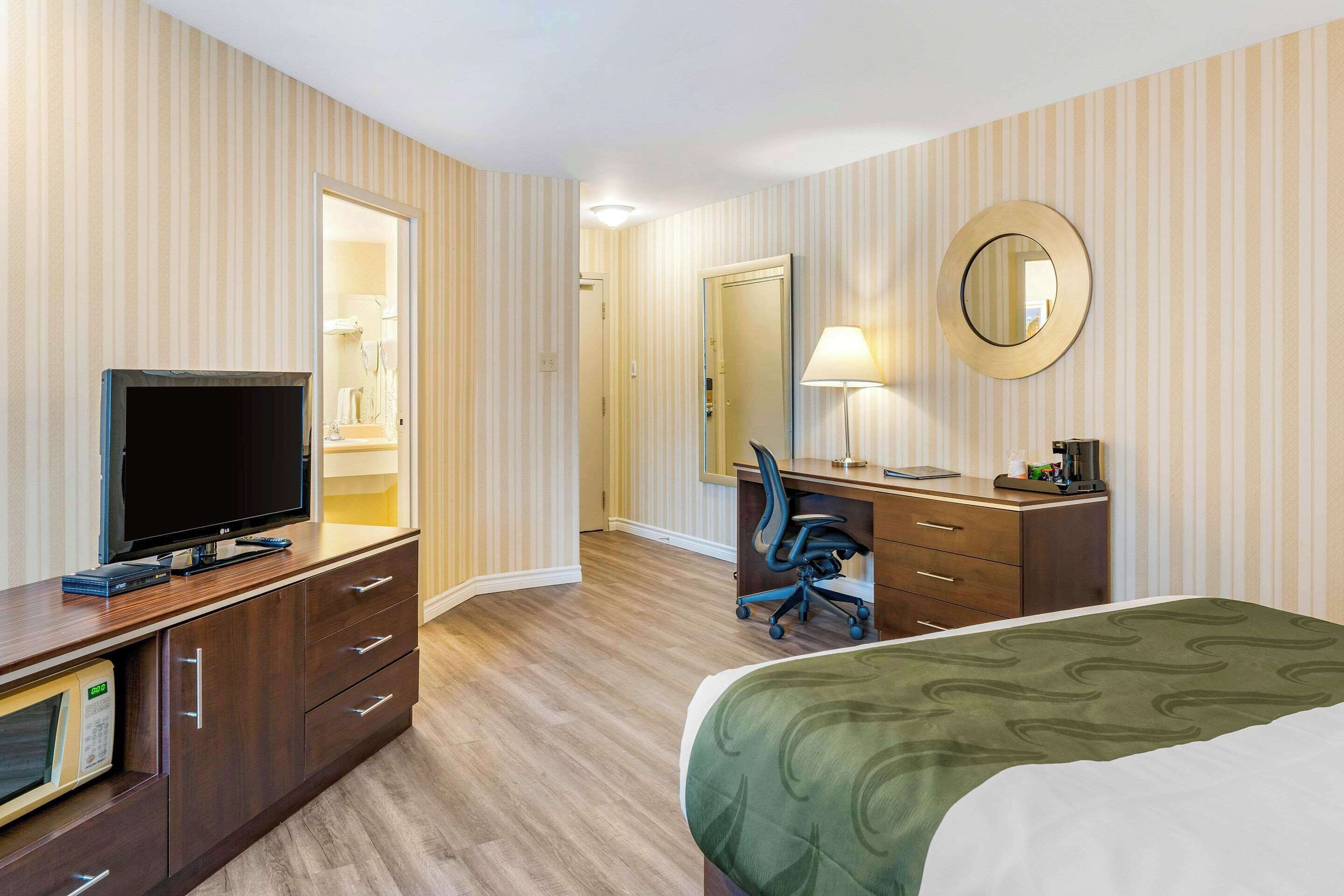 A hotel room with a flat-screen TV, a desk with a chair, a bed with a green and white bedspread, and a large mirror on the wall.