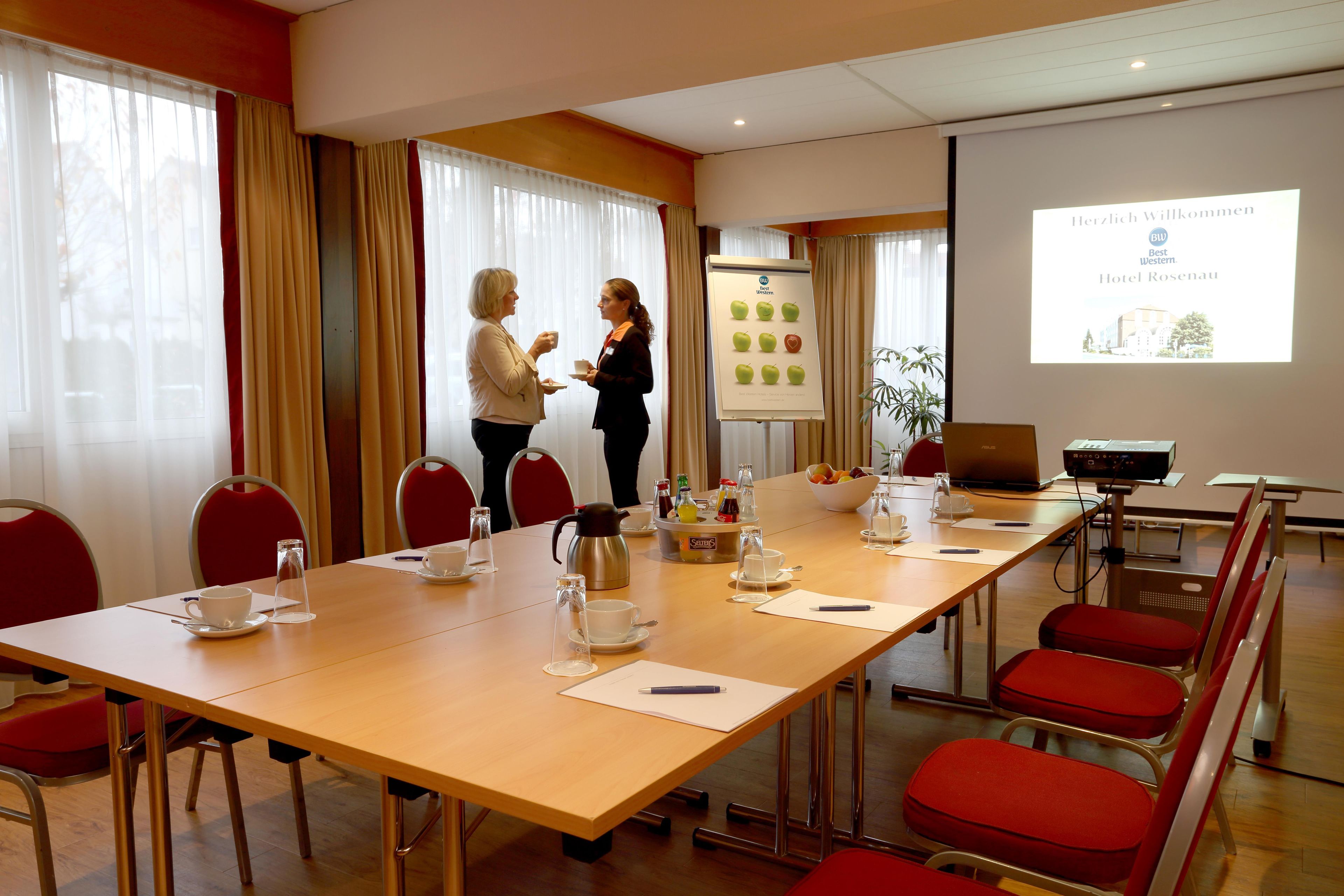 A conference room with a long table, red chairs, a projector screen, and a presentation about hotel welcome.