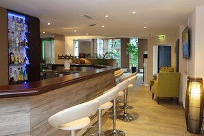 A bar area with a wooden counter and white bar stools, a seating area with a yellow chair, and a large potted plant. - Best Western Hotel Rosenau (Bad Nauheim)