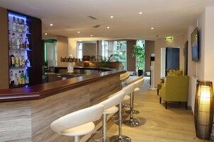 Una zona de bar con mostrador de madera y taburetes blancos, un área de asientos con una silla amarilla y una planta grande en maceta.