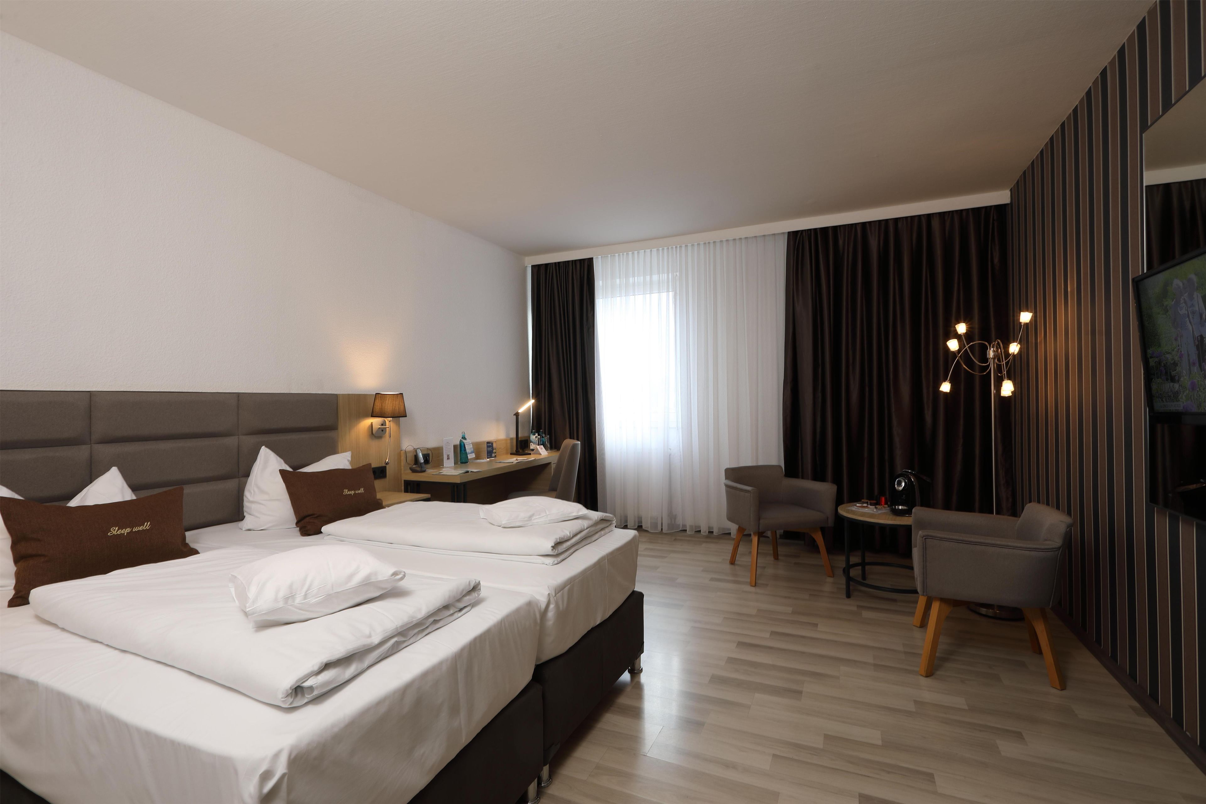 A hotel room with two beds, a desk, a chair, a TV, and a large window with curtains.