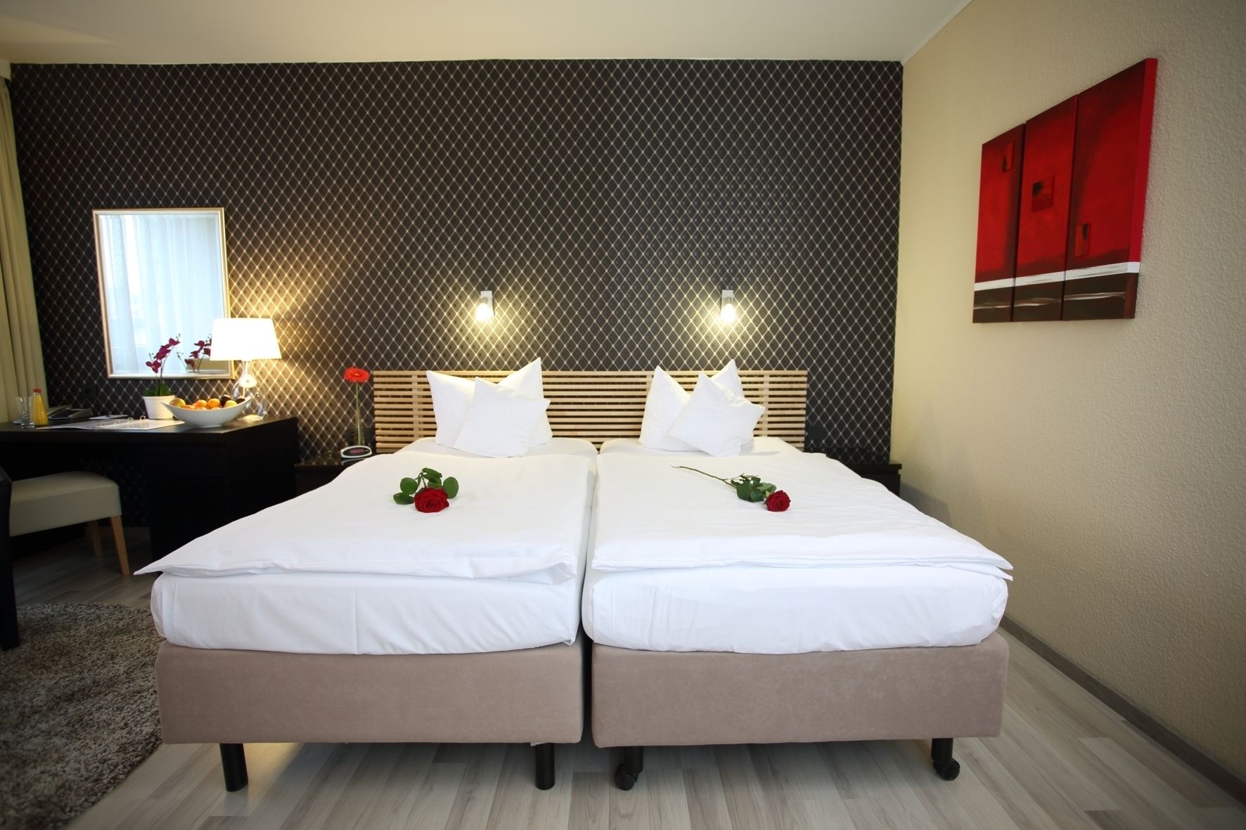 A double bed with white linens and two red roses, a bedside table with a lamp, and a desk with a chair.