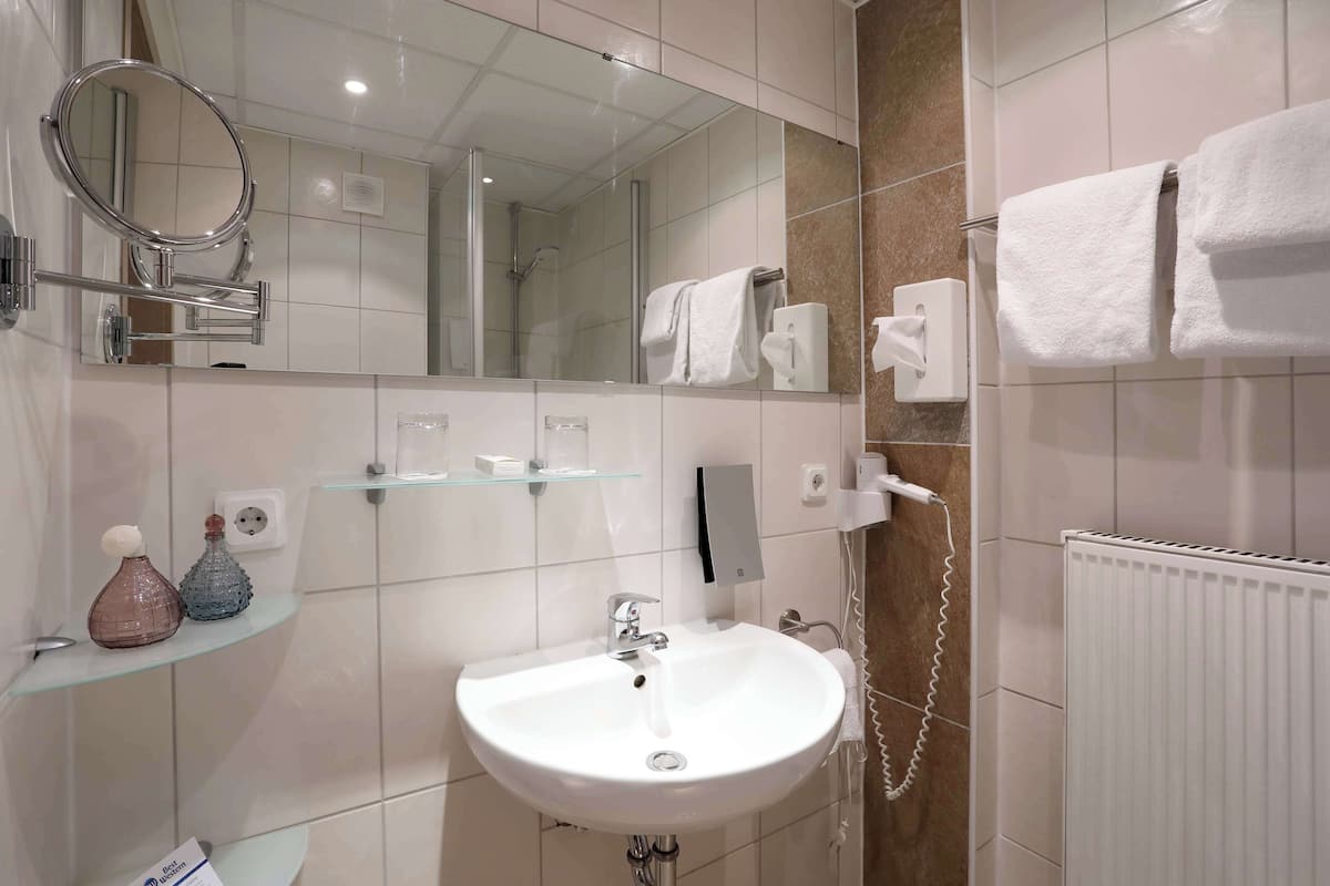 A modern bathroom with a white sink, a mirror, a glass shelf with toiletries, a towel rack, and a shower area.