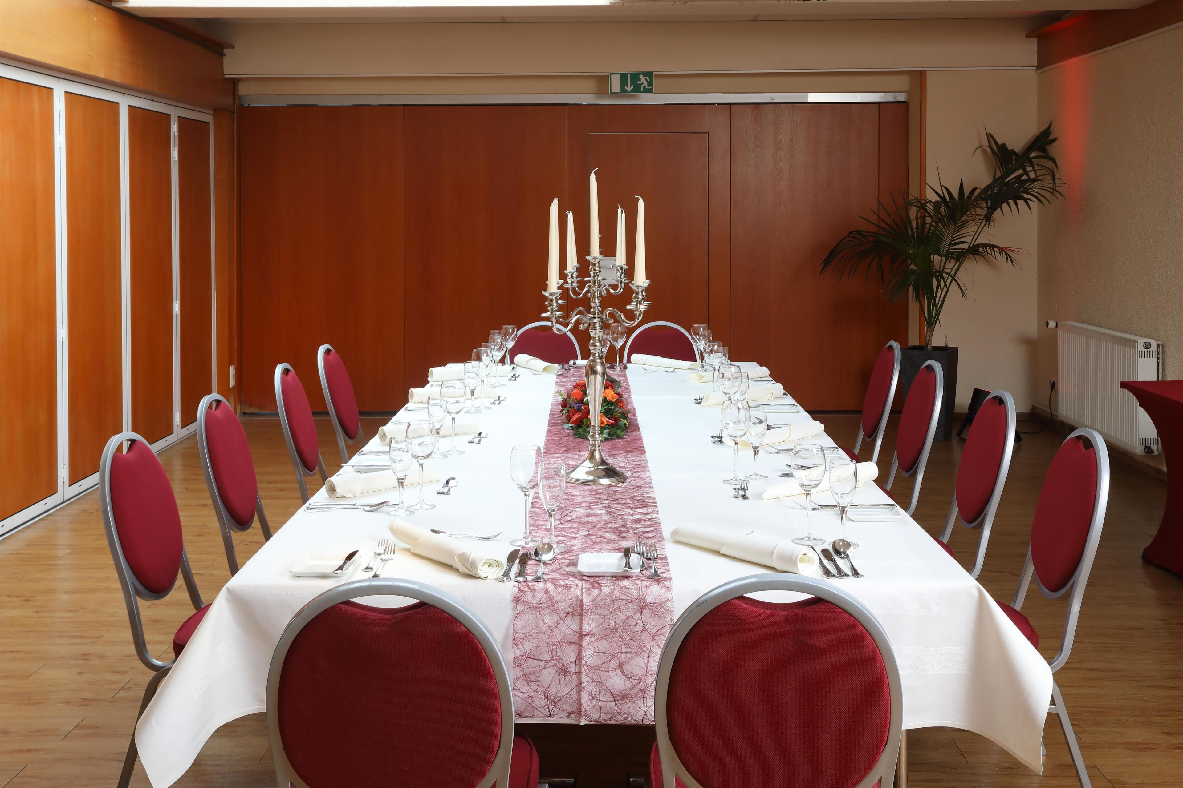 A conference room with a long table set for a formal event, equipped with chairs, a candelabra, and a potted plant.