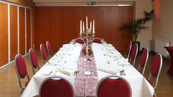 Sala de conferências com uma mesa longa posta para um evento formal, equipada com cadeiras, candelabro e planta em vaso.