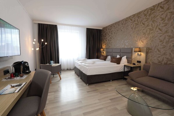 Ein Hotelzimmer mit Bett, Sofa, Schreibtisch mit Stuhl, Fernseher und einem Fenster mit Vorhängen.