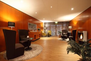 Un moderno vestíbulo de hotel con piso de madera, un mostrador de recepción, zona de estar con sillas y una pequeña mesa, y una chimenea.