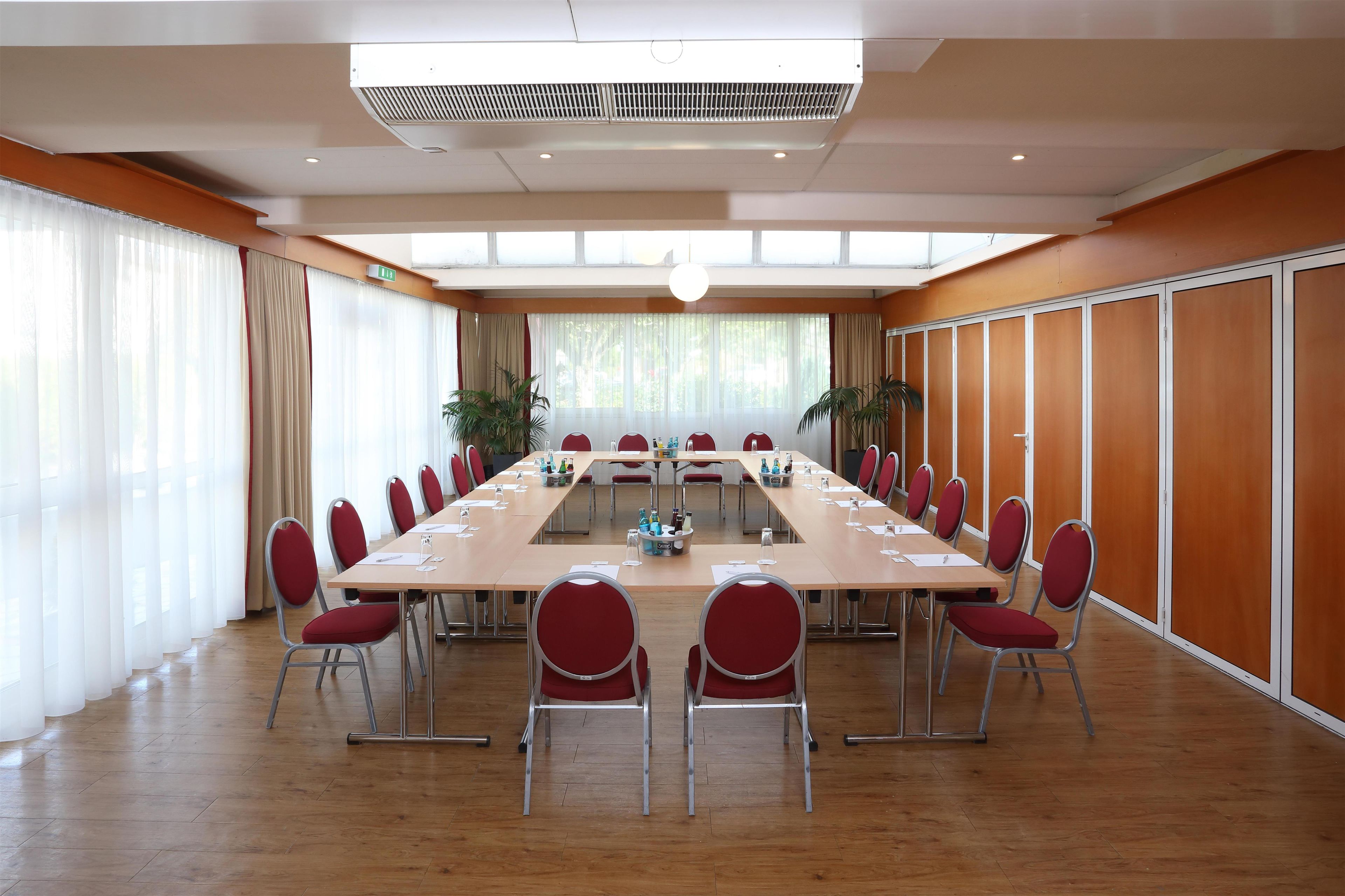 Ein Konferenzraum mit einem langen Tisch, roten Stühlen und einem großen Fenster mit Vorhängen.