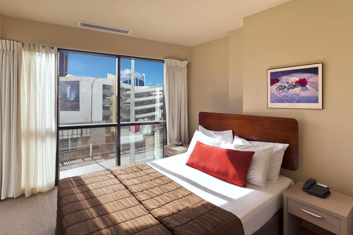 A hotel room with a large bed, a bedside table with a phone, a window with curtains, and a painting on the wall.
