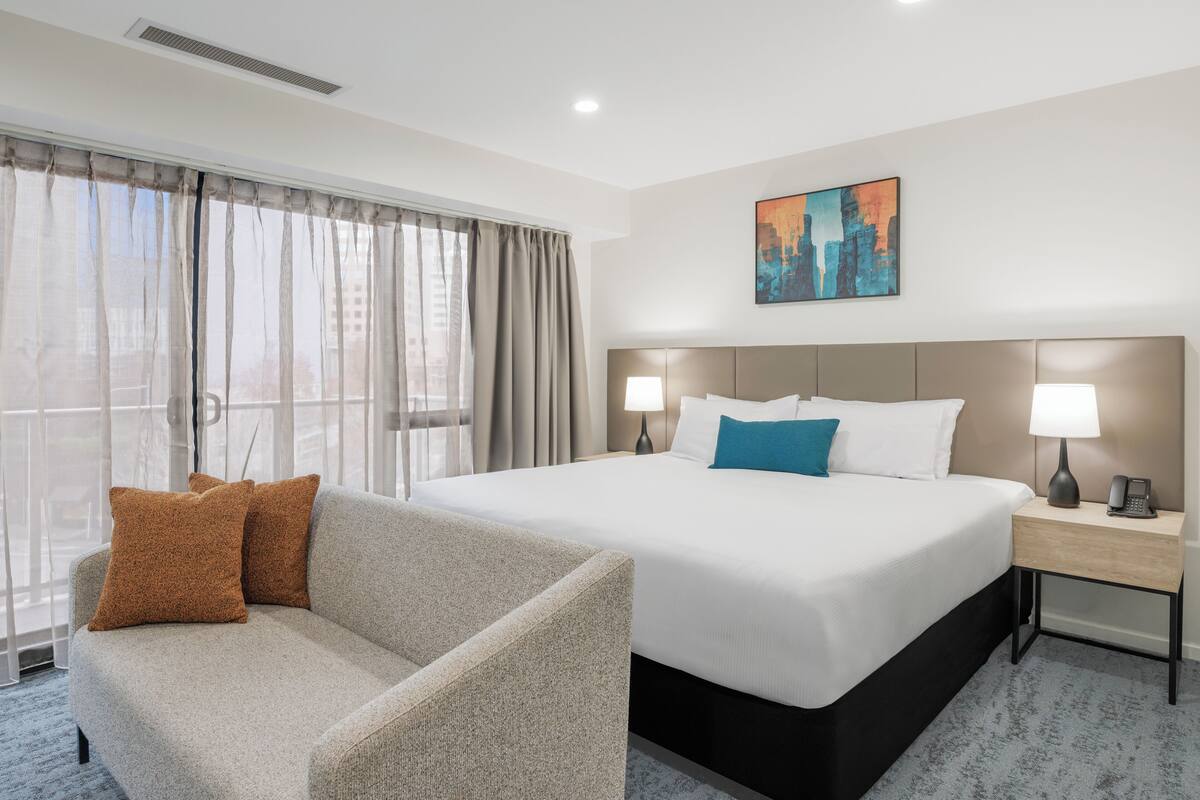 A hotel room with a bed, two bedside lamps, a nightstand, a couch, and a cityscape painting on the wall.