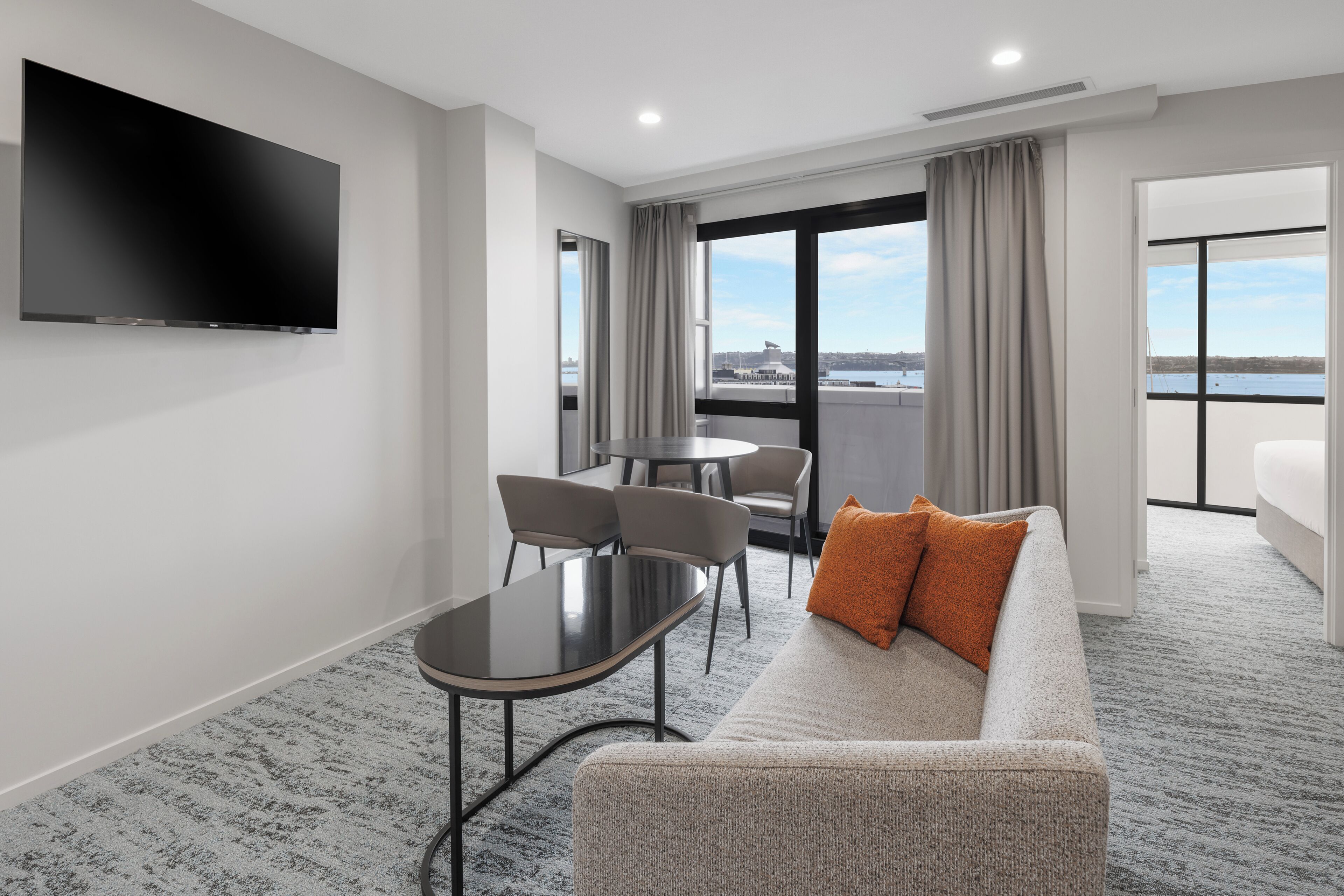 A modern hotel room with a flat-screen TV, a sofa with an orange pillow, a dining table with chairs, and a view of the city and water.