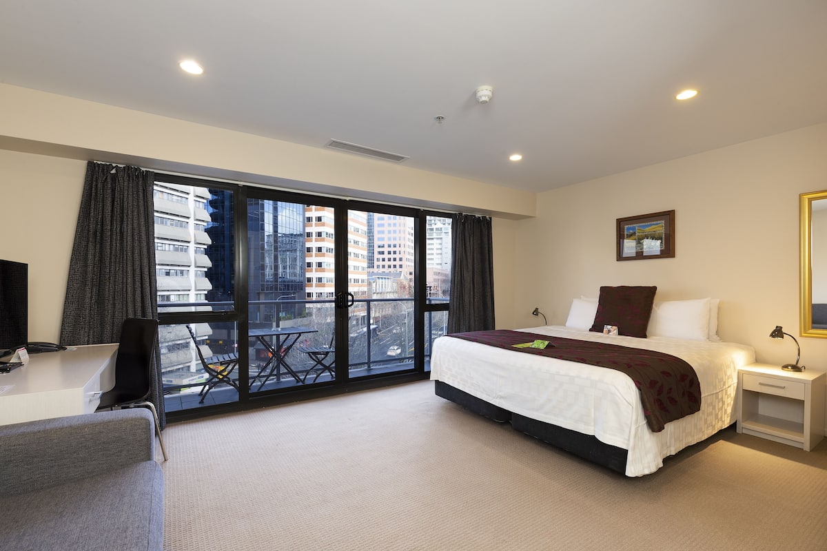 A hotel room with a bed, a desk, a chair, a small table, a nightstand, a lamp, and a window with a view of buildings.