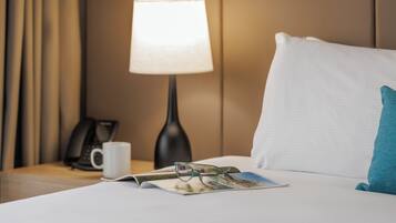 Una cama bien hecha con un almohadón, una revista, un par de gafas, una lámpara y un teléfono sobre una mesita de noche.