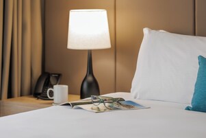 Una cama bien hecha con un almohadón, una revista, un par de gafas, una lámpara y un teléfono sobre una mesita de noche.