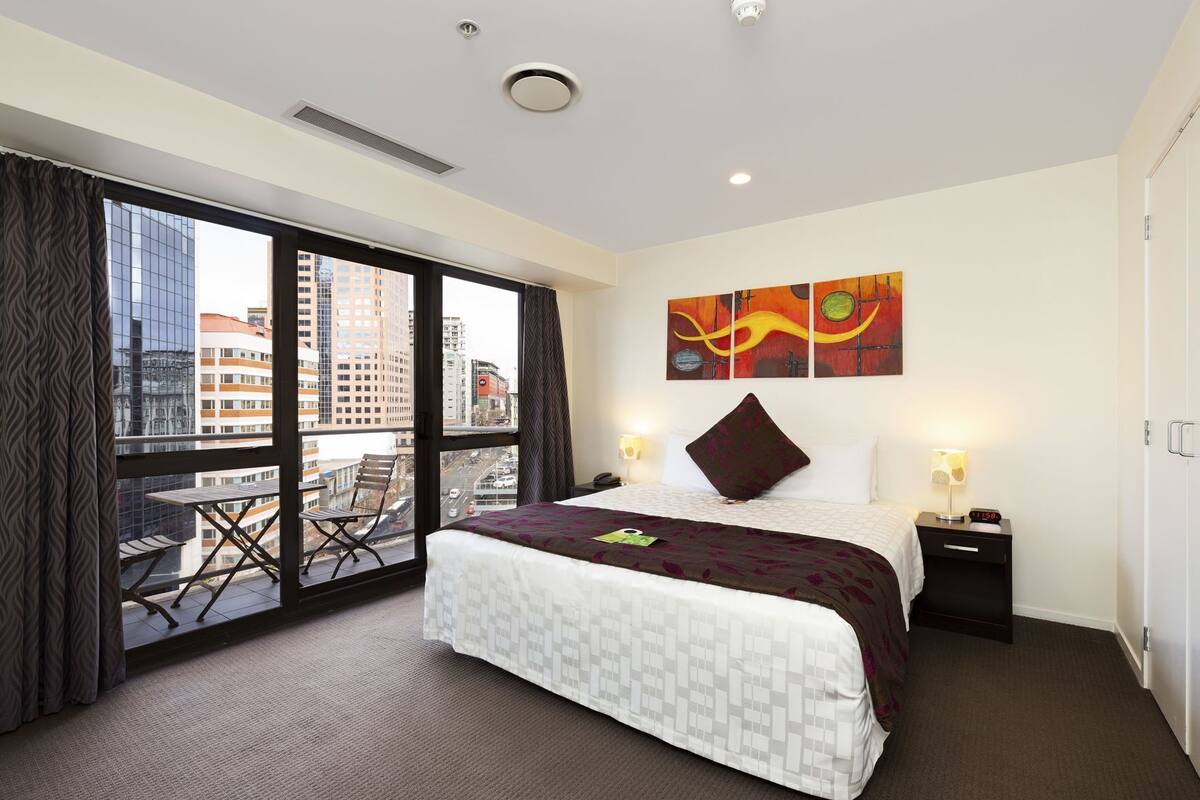 A hotel room with a bed, a nightstand, a window with a city view, and a painting on the wall.