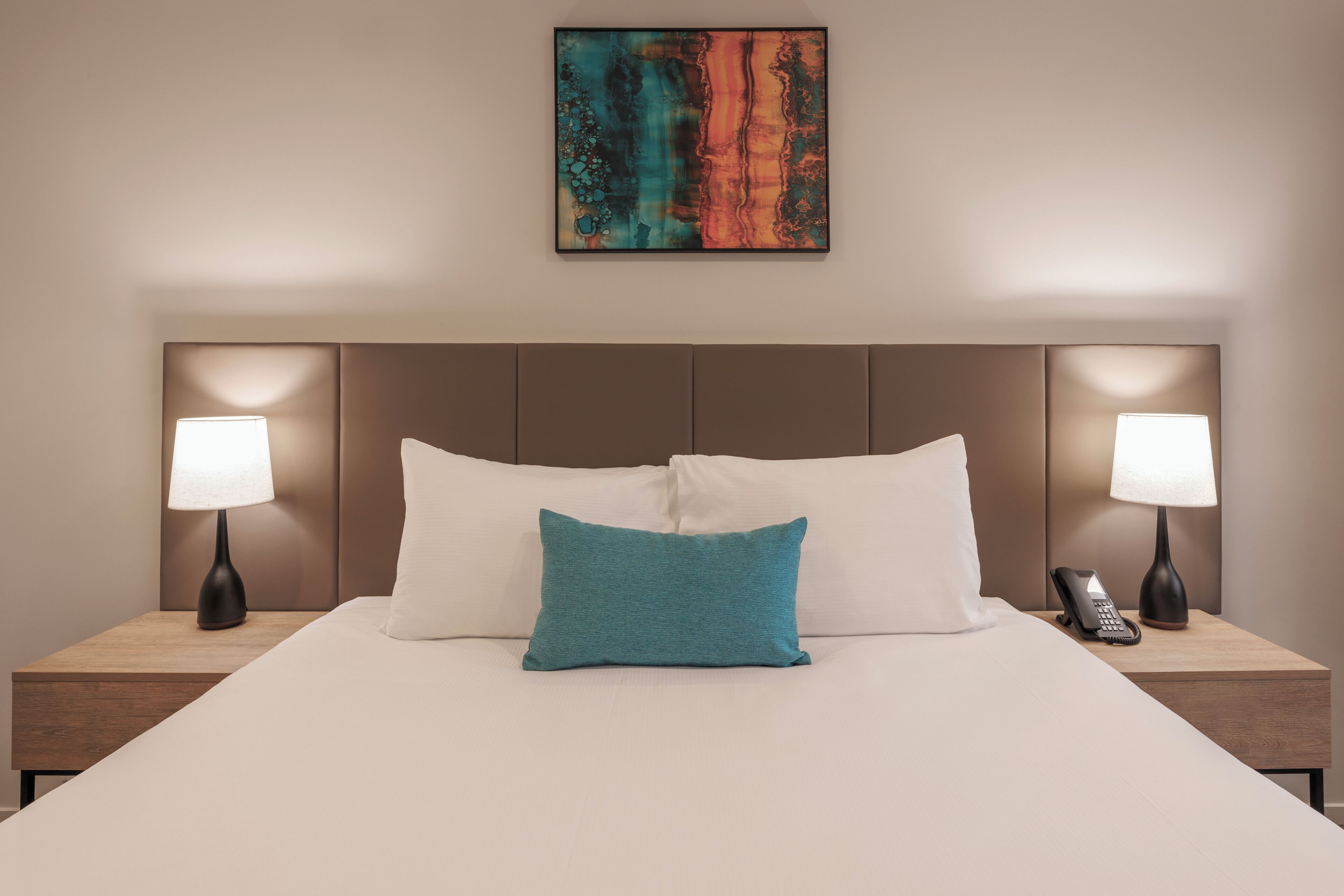 A hotel room with a large bed, two bedside lamps, a telephone, and a painting on the wall.