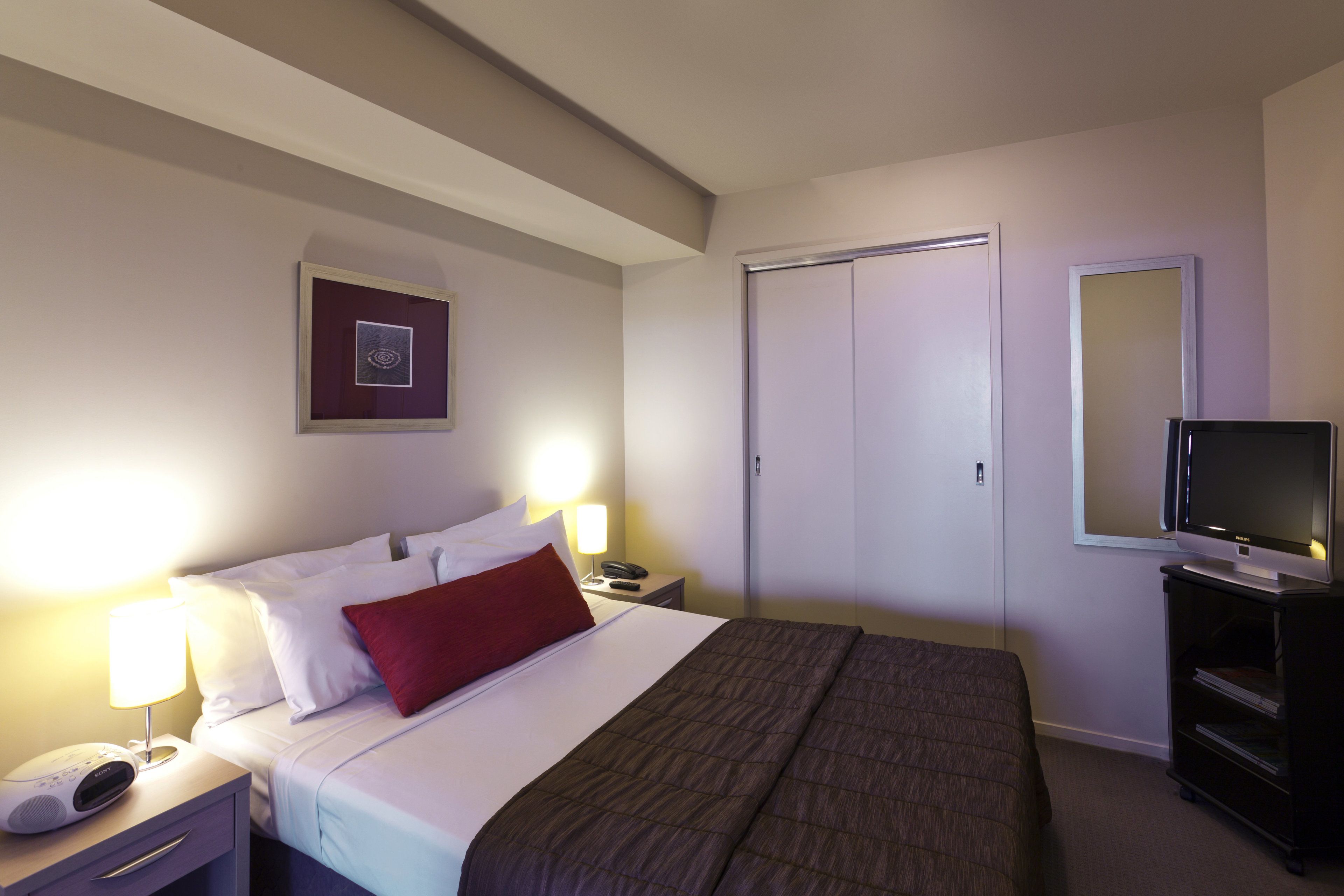 Ein Hotelzimmer mit Bett, Nachttischlampen, Fernseher, Kleiderschrank und einem gerahmten Bild an der Wand.