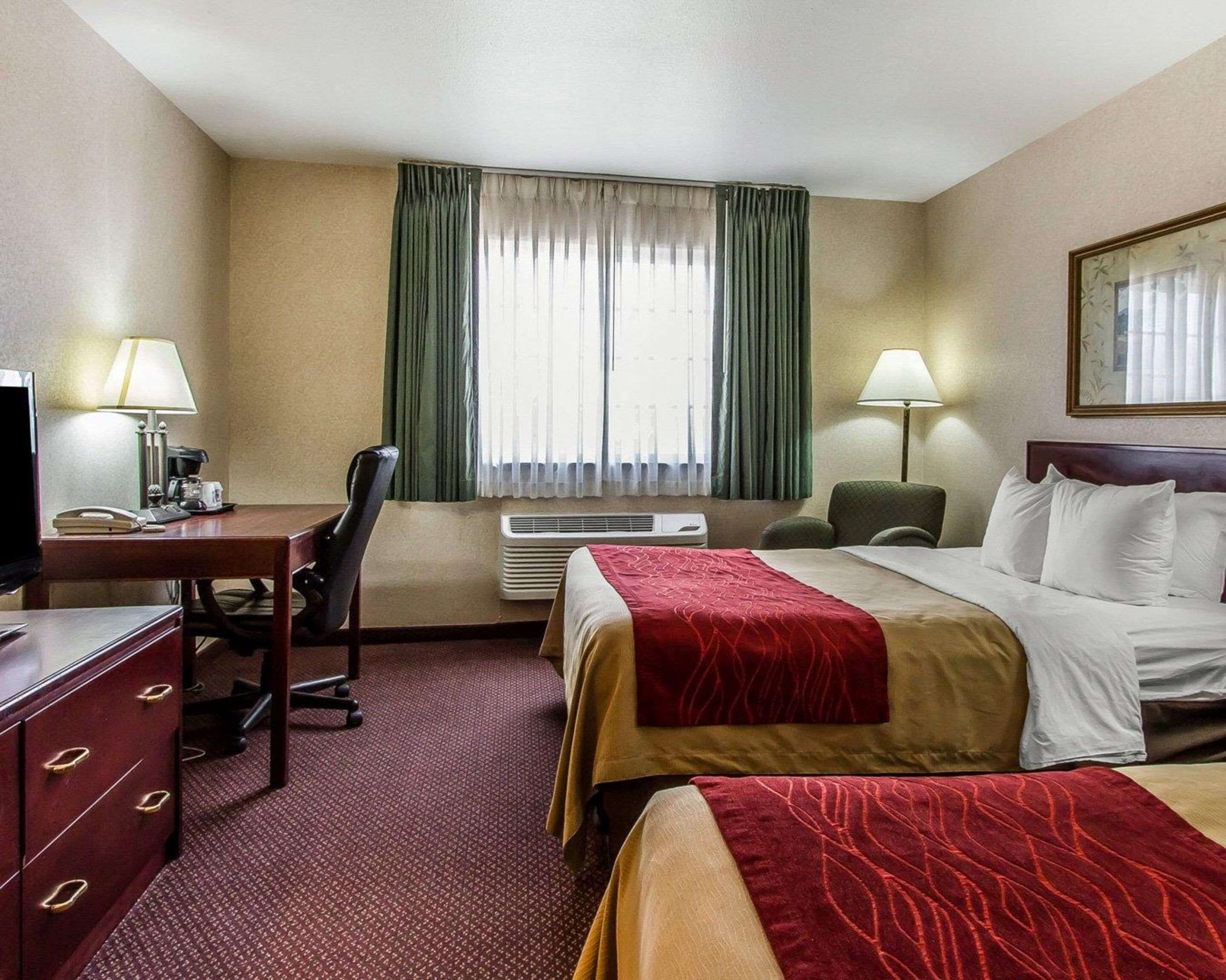 Una habitación de hotel con cama, escritorio con teléfono, silla, lámpara y ventana con cortinas.