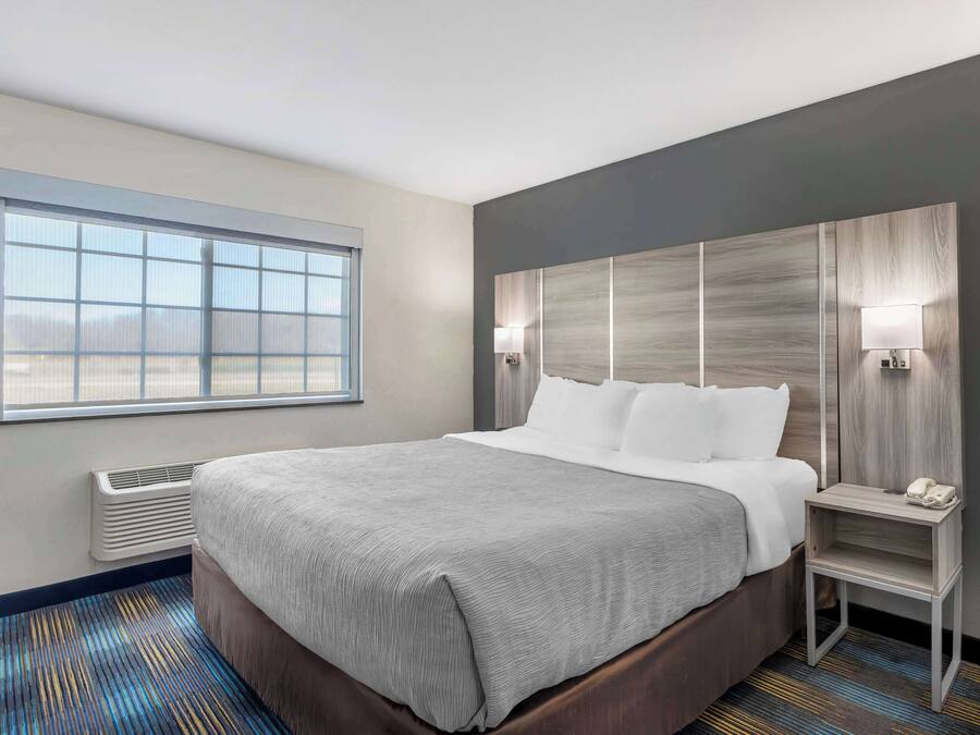 A hotel room with a large bed, a nightstand with a phone, a window with blinds, and a wall-mounted heater.