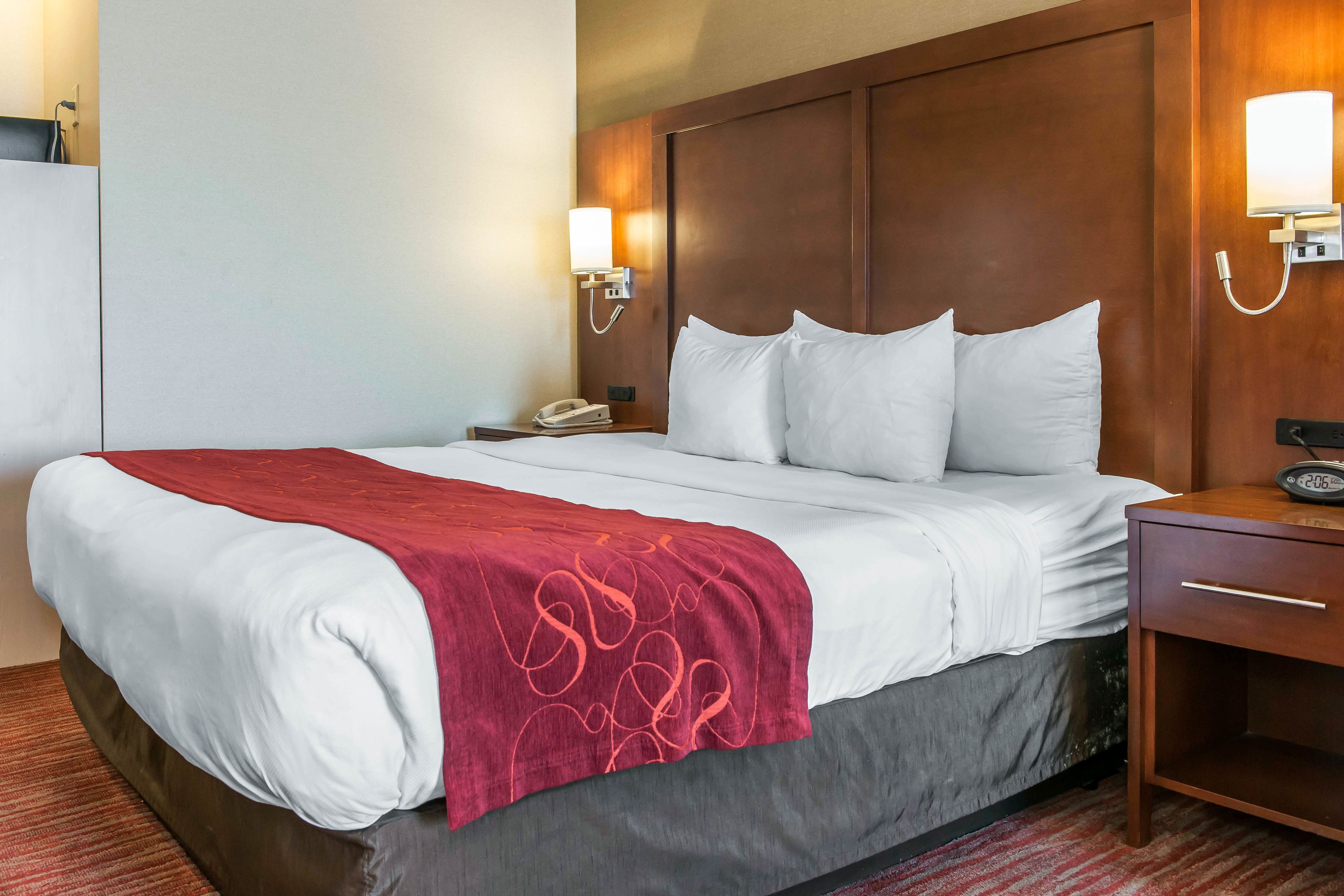 A hotel room with a large bed, a wooden headboard, two bedside lamps, a nightstand with a phone, and a red throw blanket.