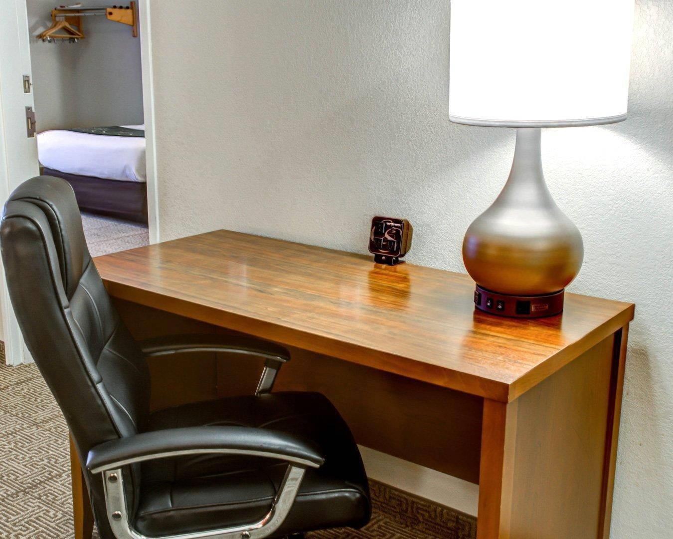 A hotel room with a wooden desk, a black leather chair, a bedside table, a lamp, and a small clock.