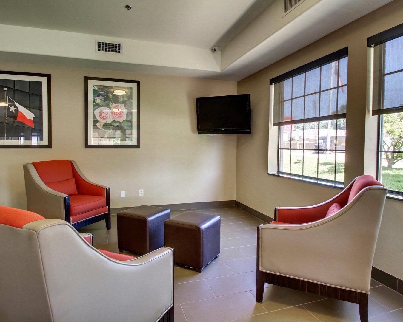 A room with a flat-screen TV mounted on the wall, two framed floral paintings, and a seating area with three chairs and a small ottoman.