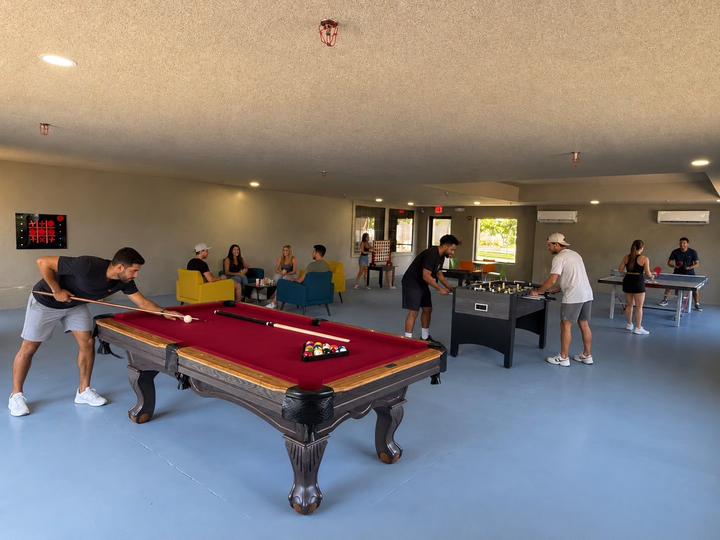A pool table in a room with a blue floor, a foosball table, and a group of people playing pool.