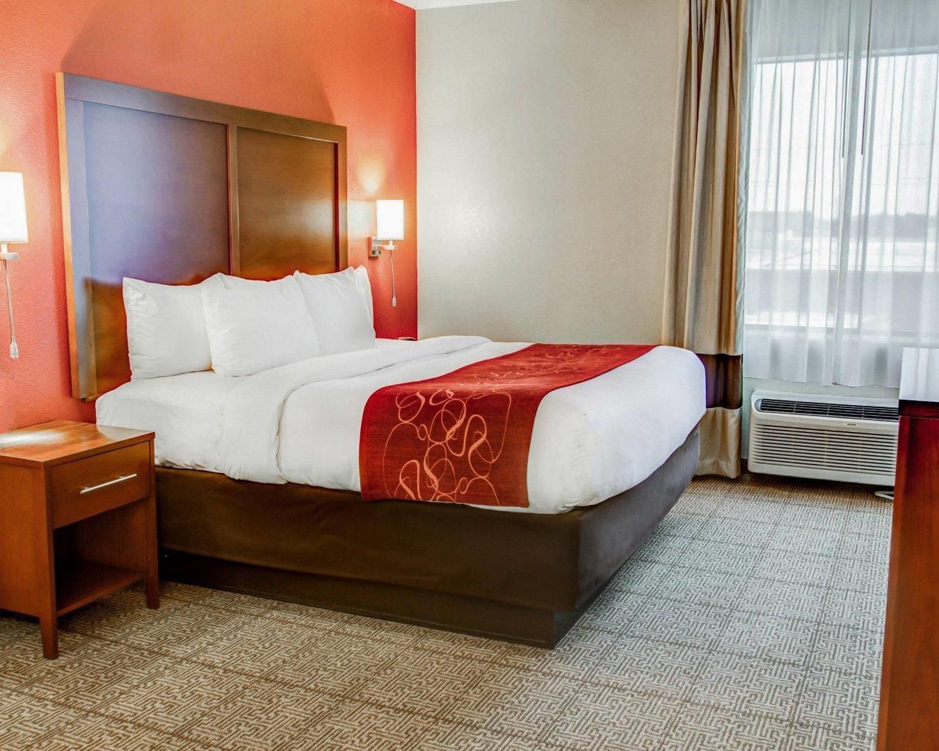 A hotel room with a large bed, a nightstand, a wall-mounted lamp, and a window with curtains.