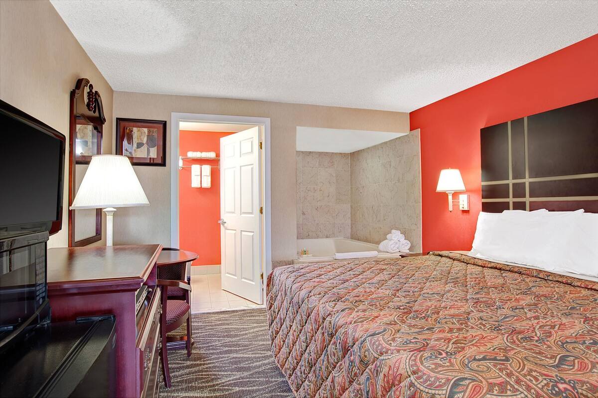 Une chambre d’hôtel avec un lit, un bureau doté d’une lampe, une télévision et une salle de bain visible à travers une porte ouverte.
