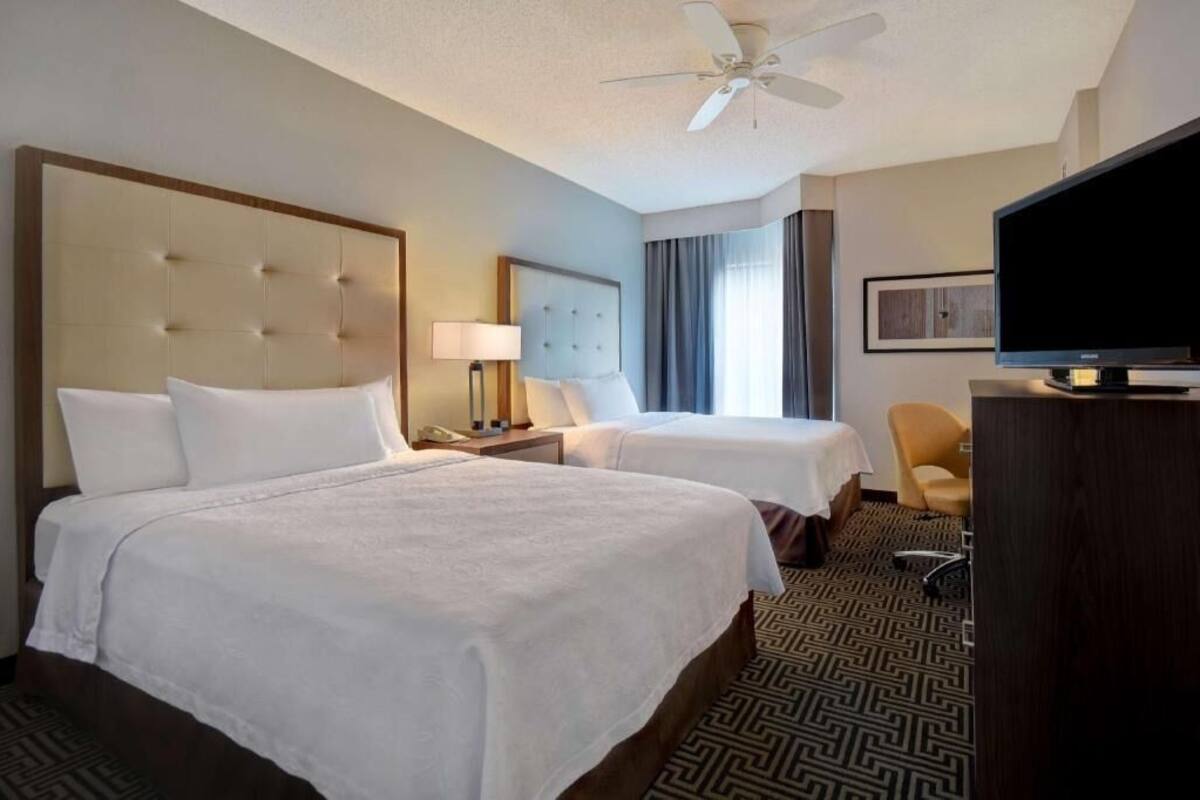 A hotel room with two beds, a television, a ceiling fan, and a desk with a chair.