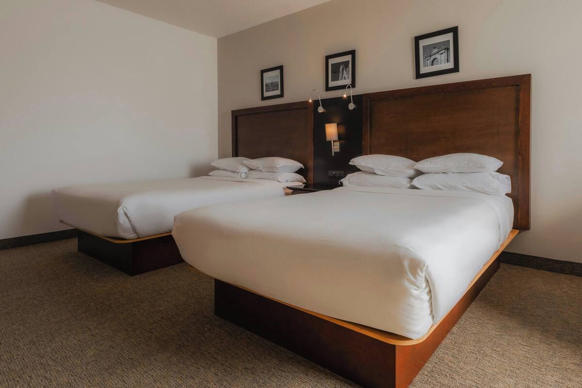 Habitación de hotel con dos camas, cabecero de madera, mesita de noche y una lámpara de pared.
