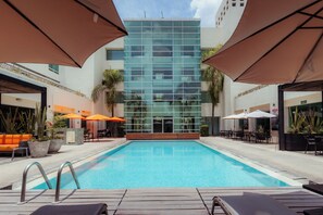 A swimming pool with lounge chairs and umbrellas, surrounded by a building with large windows. - Sheraton Leon (León)