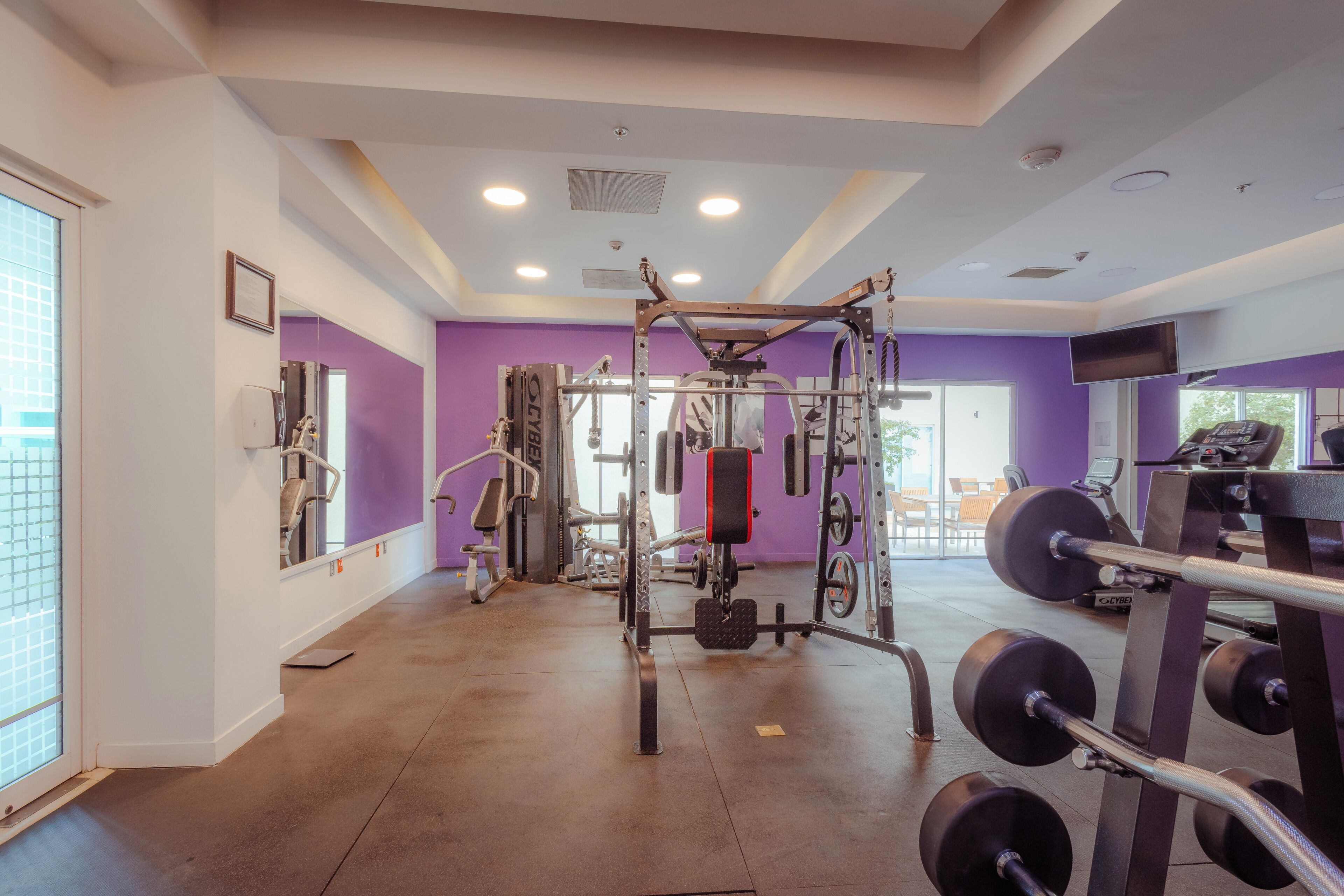 Ein Fitnessraum mit unterschiedlichen Trainingsgeräten, einer violetten Wand und einem Fenster.