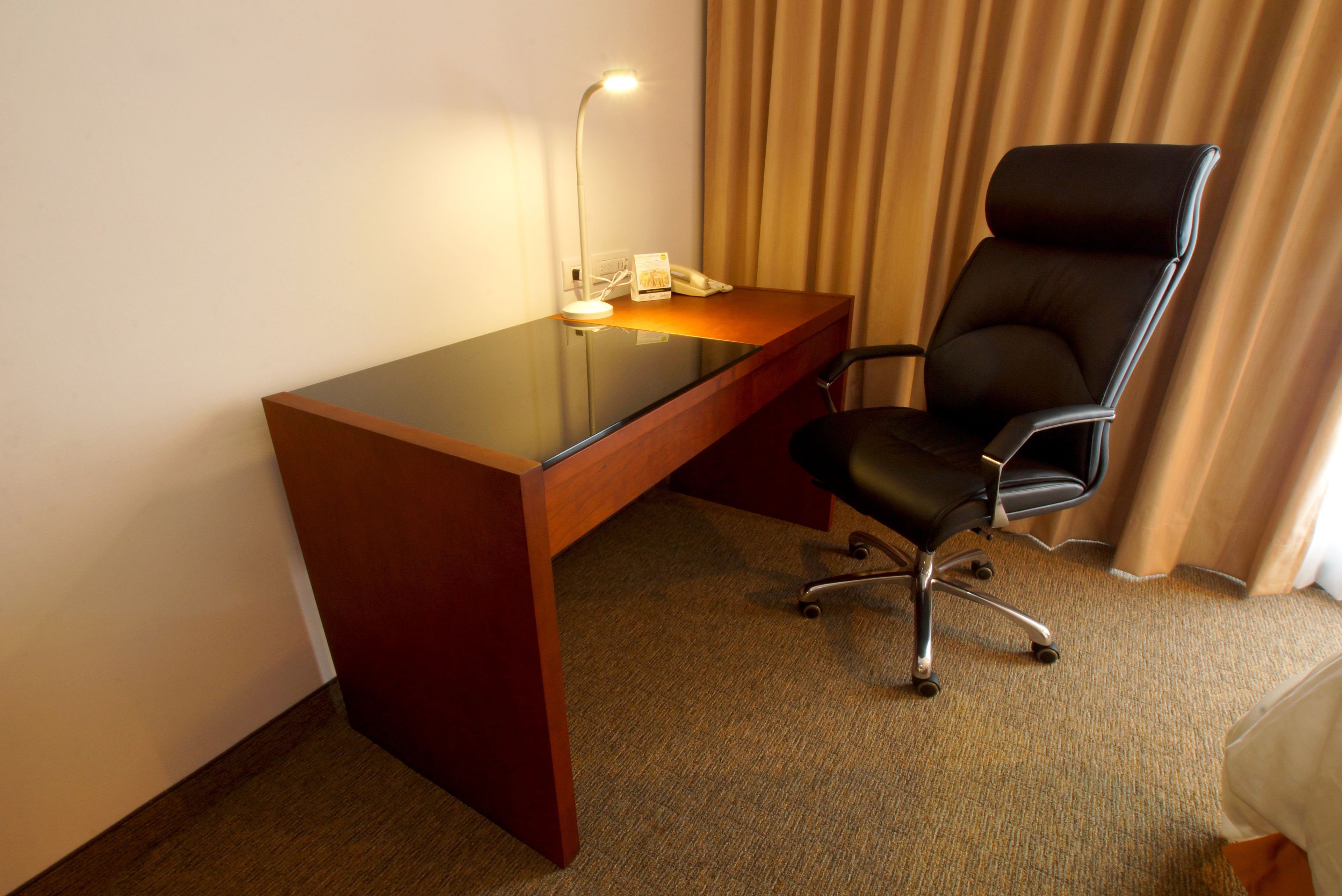A hotel room with a wooden desk, a black office chair, a desk lamp, and a telephone.