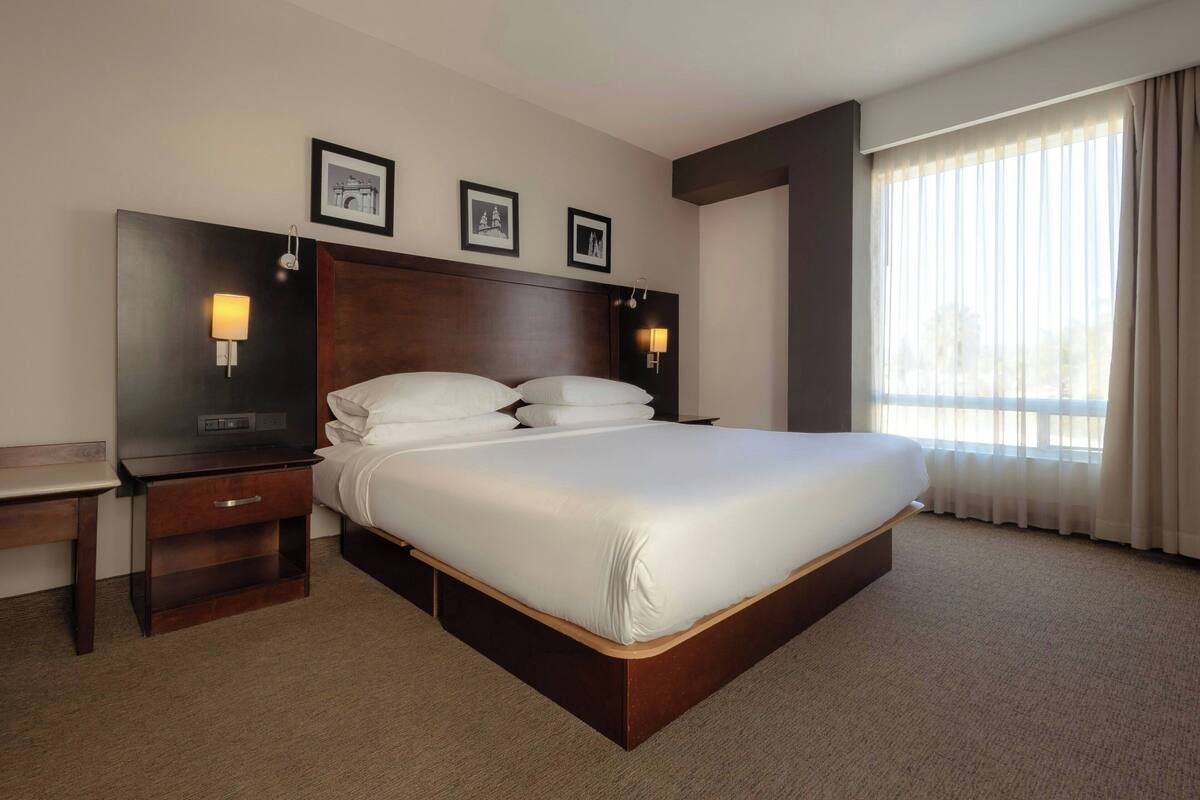 Habitación de hotel con una cama grande, dos mesitas de noche, cabecero y un amplio ventanal con cortinas.